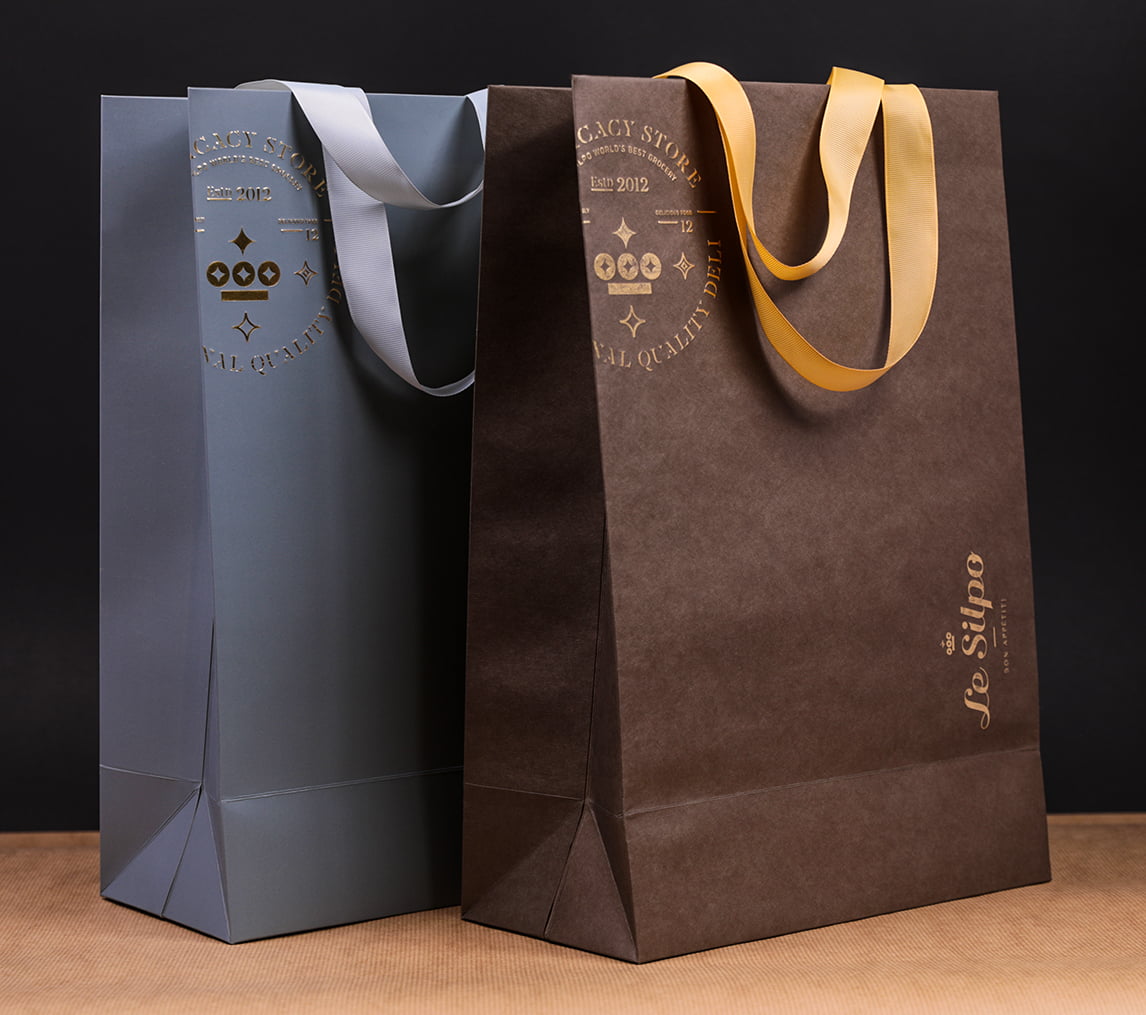 Картонні пакети «Le Silpo» - особливі тим, що два принципово різні стилі: еко-пакет з крафт паперу  та ламінований пакет з картону ми поєднали в одну лінійку з допомогою вишуканого тиснення золотою фольгою та конгреву. Матова ламінація доповнює неповторність та вишуканість сірого кольору. Світло- сірі широкі атласні ручки, приємні на дотик і міцно закріплені у спеціально вирізаних отворах гармонійно підійшли обом стилям.